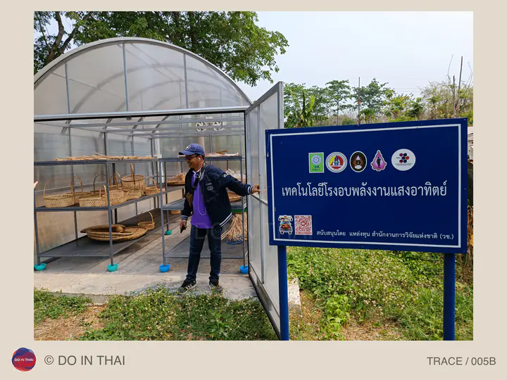 A person stands beside a transparent solar drying house with trays inside and a Thai sign explaining the technology.