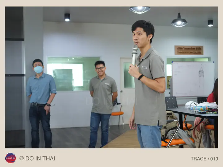 A team member speaks into a microphone in a workroom with colleagues behind him.