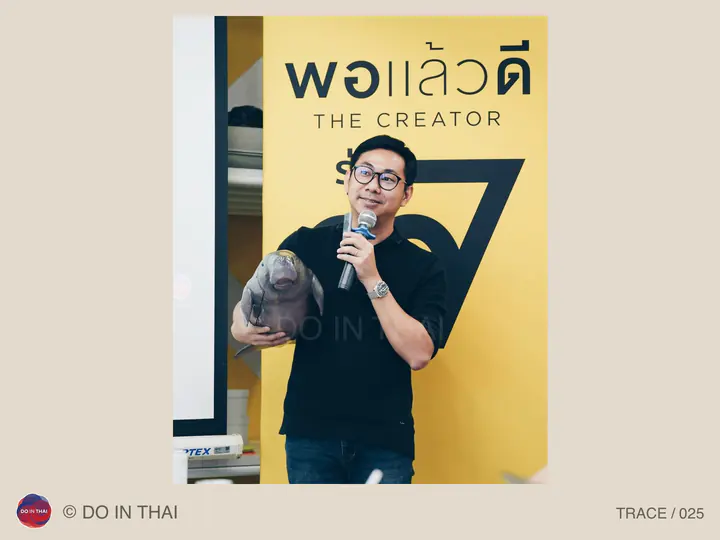 A speaker holds a microphone and an animal-shaped object in front of a Por Laew Dee banner.