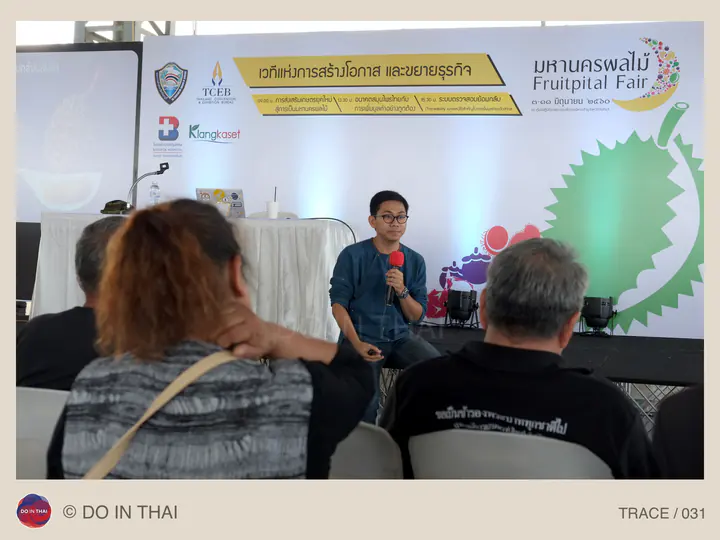 A speaker holds a red microphone in front of a fruit fair banner, with listeners nearby.