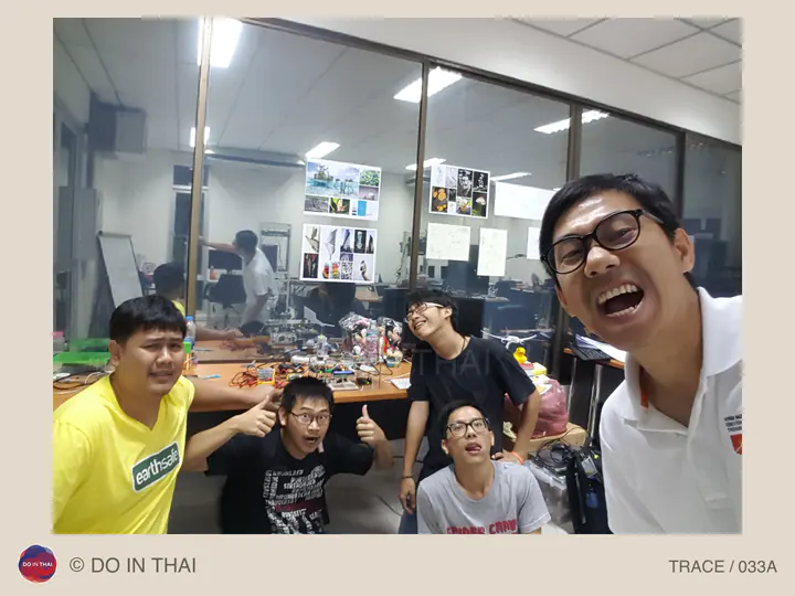 A group takes a playful selfie in a workroom, with electronics on the table and photographs on a glass partition.