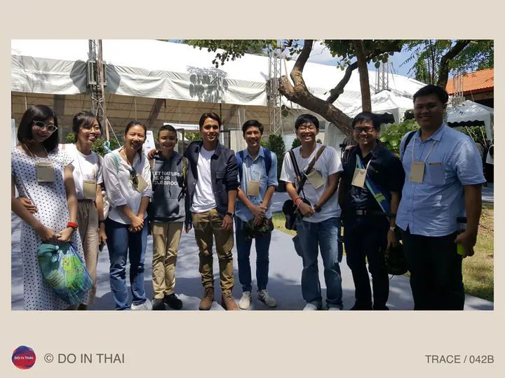 A group wearing event badges poses outdoors beside white tents and trees in bright sunlight.