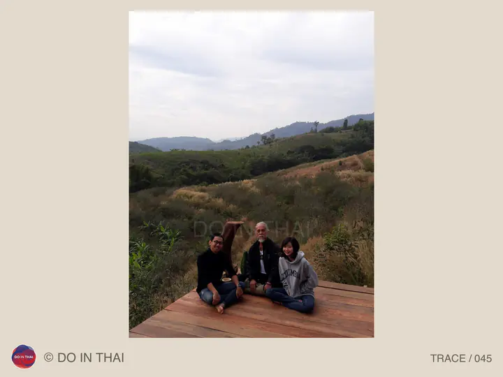 Three people sit on a wooden deck overlooking fields and distant hills.